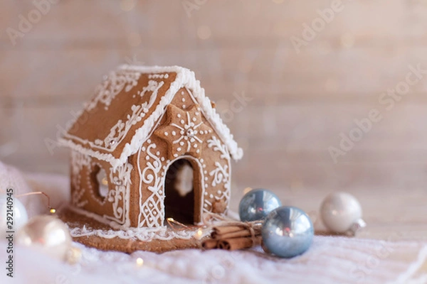 Fototapeta Gingerbread house, Christmas cozy decorations on wooden and knitted background with glares. Handmade sweets is decorated with holly, blue and white ornaments, new year lights, garland.