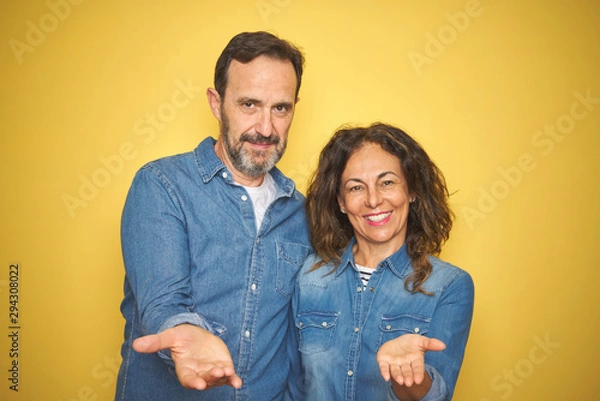 Obraz Beautiful middle age couple together standing over isolated yellow background smiling cheerful offering palm hand giving assistance and acceptance.