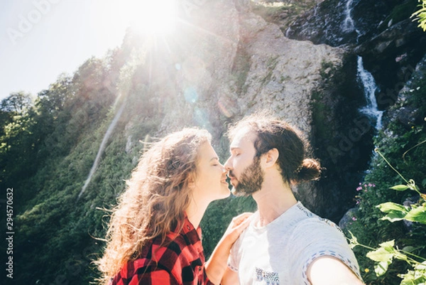 Fototapeta Young happy couple enjoing the waterfall. Couple standing in front of waterfall and kissing. Concept of happy travel together.