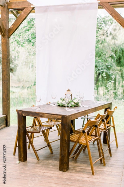 Fototapeta Wedding. Banquet. The wooden chairs and table for guests, decorated with flowers and greenery, served with cutlery and wineglasses. The table stands on a wooden floor in reastaurant outdoors in forest