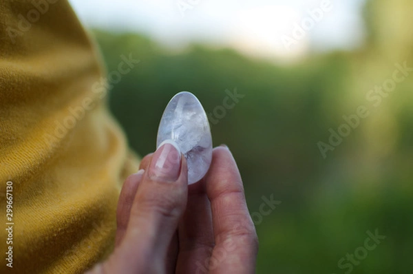 Fototapeta Adult girl in yellow shirt and blue jeans is holding transparent violet amethyst yoni egg for vumfit, imbuilding or meditation outdoors on her body background outdoors