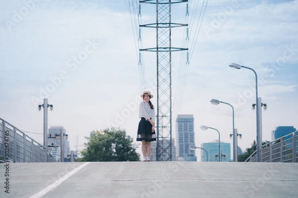 Fototapeta Cheerful pretty asian girl in white shirt black skirt and boater hat standing in the center slope of green pathway and touching her arms her face is full of happiness on a beautiful blue sky day