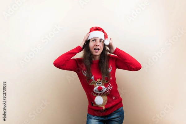 Fototapeta Young woman in Christmas knitted sweater with reindeer and Santa's hat holding head on beige background