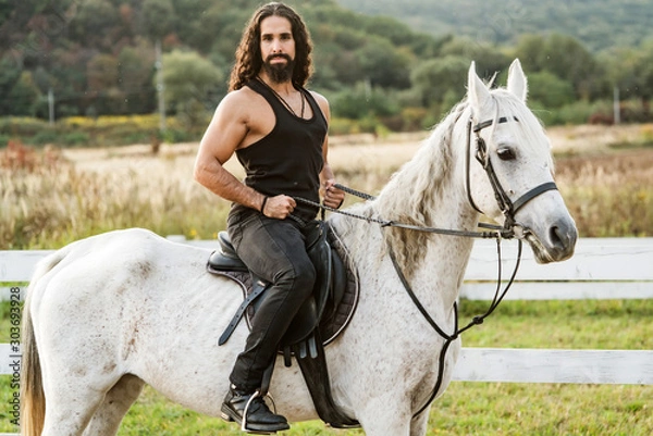 Obraz Young jockey training his horse for a ride. Good looking, hunky cowboy rides horse at beautiful country side nature. Equestrian rides his white well looking horse.