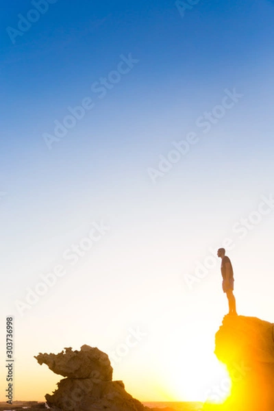 Fototapeta man standing on a rocky mountain cliff at golden hour wearing a black t-shirt and shorts with the golden sun rays peaking from underneath the rocks 