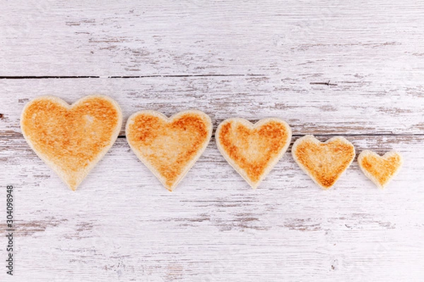 Fototapeta Group of toasted bread hearts one each smaller together in a row on white wooden table background. The concept of support in big family.