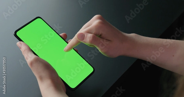 Fototapeta Young woman sitting on a chair near window and using smartphone with green screen