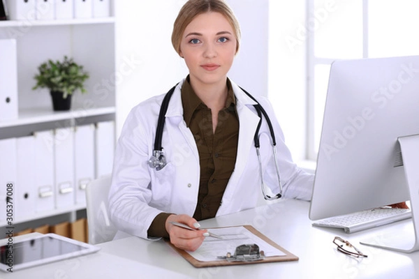 Fototapeta Young woman doctor at work in hospital looking at desktop pc monitor. Physician controls medication history records and exam results. Medicine and healthcare concept