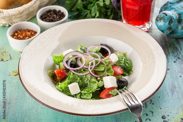 Fototapeta Fresh Greek salad with olives, tomato, feta cheese, cucumber and onion and glass of red drink on blue wooden table