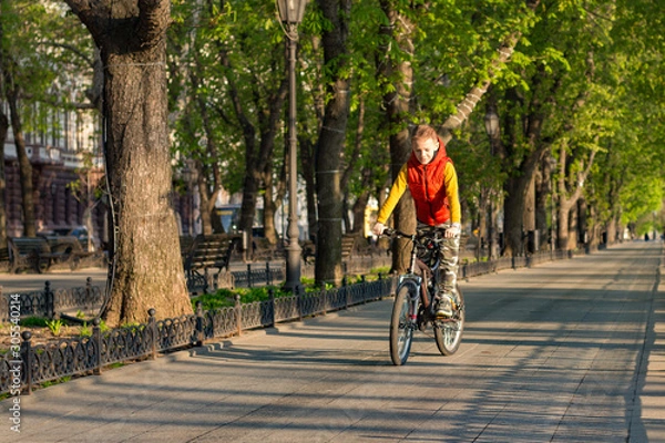 Fototapeta A boy of 8 years old in an orange vest rides a bicycle in a city park on a sunny spring morning.