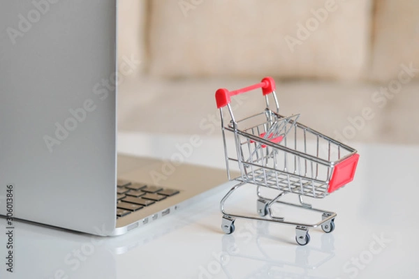 Fototapeta A gray laptop on a table next to a shopping cart from a supermarket. Close up.