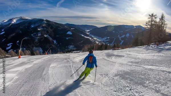 Fototapeta A man with a small backpack skiing on a perfectly groomed slopes in Bad Kleinkirchheim, Austria. There are lots of snow caped mountains around him. Few trees on the side. Winter sports.