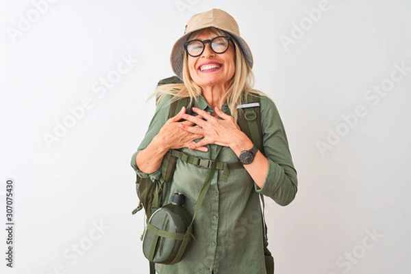 Fototapeta Middle age hiker woman wearing backpack hat canteen glasses over isolated white background smiling with hands on chest with closed eyes and grateful gesture on face. Health concept.