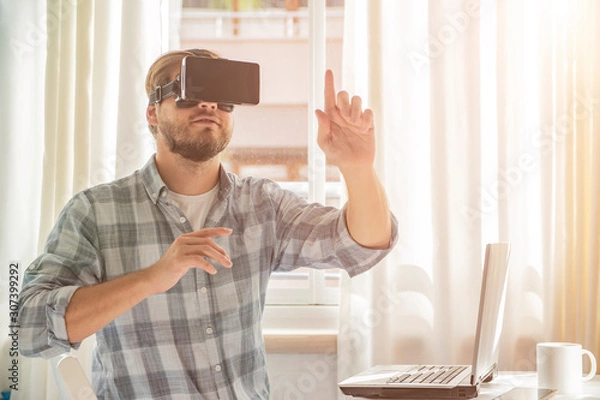 Fototapeta  Young bearded man wearing virtual reality glasses in modern interior,    blurred background. Handsome caucasian male using VR headset, playing   video games.