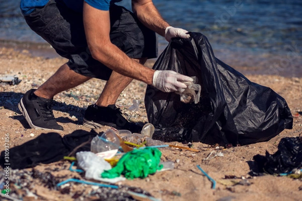 Fototapeta Man with black bag collect garbage on the beach. Environmental pollution concept