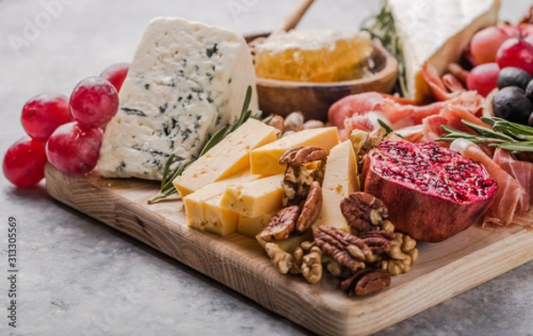 Fototapeta Traditional italian antipasto plate. Assorted cheeses on wooden cutting board. Brie cheese, cheddar slices, gogonzola, walnuts grapes, olives, prosciutto, rosemary and glass of red wine. top view