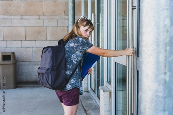 Fototapeta Sad/depressed teen girl/student opening a glass door to her school while wearing a backpack and holding binders.