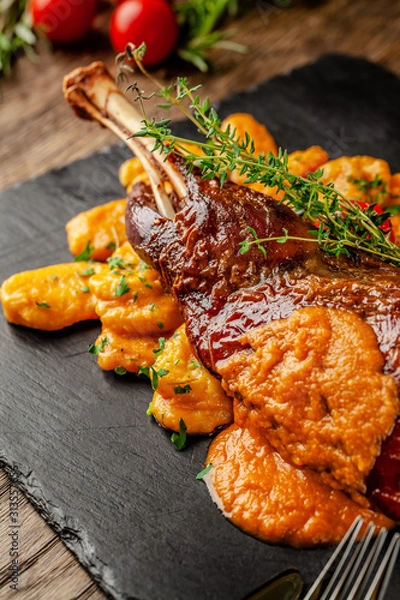 Fototapeta Serbian, Balkan cuisine. Lamb of leg with gnocchi. Serving in a restaurant on a black slate, on wooden table. background image, copy space