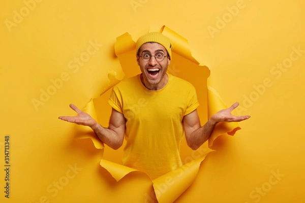 Fototapeta Horizontal shot of friendly looking happy Caucasian man spreads hands sideways, presents something cool, dressed in casual clothes, poses in paper hole alone, has good attitude, advertises item