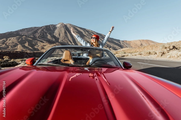 Fototapeta Woman traveling by convertible car on the picturesquare volcanic valley. Wide view with red car hood on the foreground