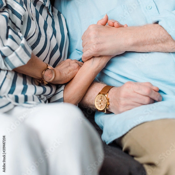 Fototapeta Cropped view of senior man holding hand of wife on couch