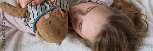 Fototapeta Pretty little girl is hugging teddy bear while sleeping in bed at home