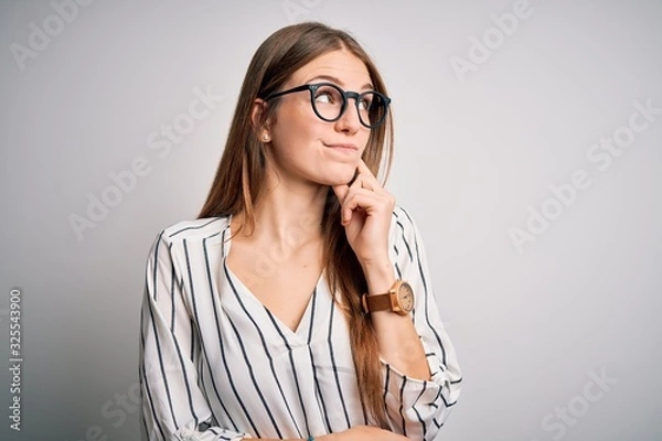 Fototapeta Young beautiful redhead woman wearing casual striped t-shirt and glasses with hand on chin thinking about question, pensive expression. Smiling with thoughtful face. Doubt concept.