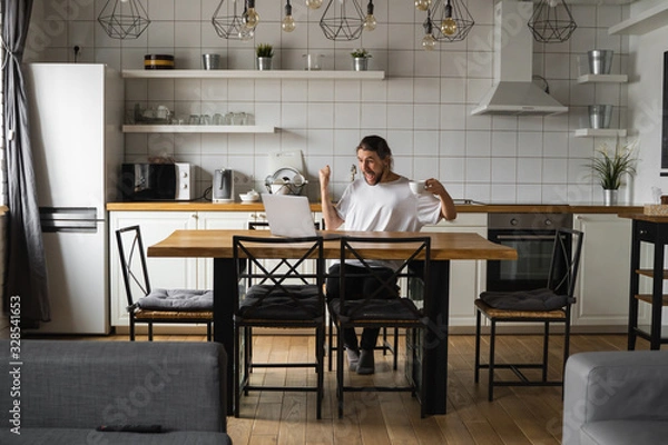 Fototapeta Young freelancer succeed with project working from home. Excited glad young freelancer triumphing with raised fists. Euphoric winner happy man using a laptop in a desk at home. Successful bearded man.