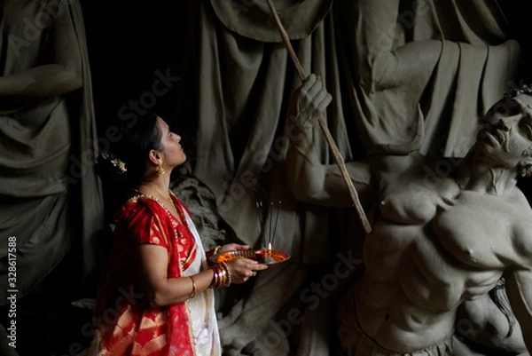 Fototapeta An young and beautiful Indian Bengali brunette woman in red white traditional ethnic sari holding a puja thali for worshiping goddess Durga clay idol . Indian culture, religion and fashion