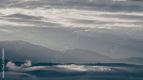 Fototapeta clouds and rays of dawn in mountain valley, soft light in early morning, meditation in nature. Gentle hills in bluish haze, silhouettes of mountains