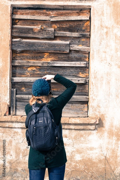 Fototapeta Girl photographer with a backpack, on the street in a green hat and jacket photographs a clogged (boarded up) board with an old window. Back view.