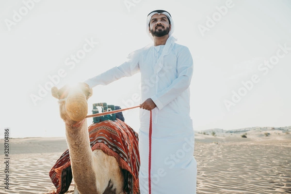 Fototapeta Man wearing traditional clothes, taking a camel out on the desert sand, in Dubai