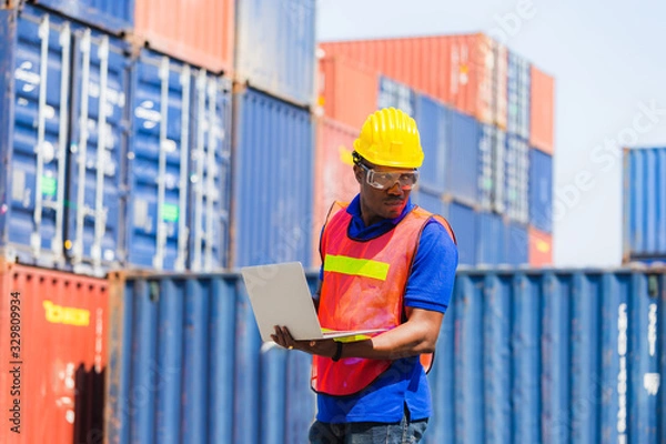 Obraz Worker man in hardhat and safety vest holding laptop, Foreman control loading containers box from cargo