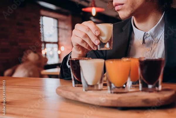 Obraz young curly-haired guy in a black suit drinking a liqueur in the bar