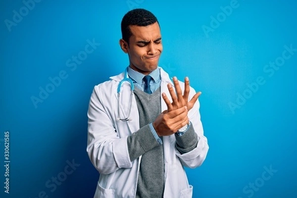 Fototapeta Handsome african american doctor man wearing coat and stethoscope over blue background Suffering pain on hands and fingers, arthritis inflammation