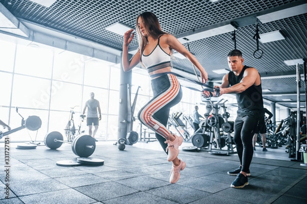 Fototapeta Young sportive people doing exercises together in the gym. Woman running when man holding her by elastic tape