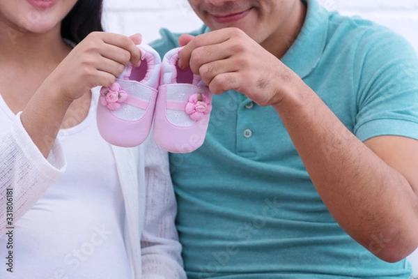 Obraz Close up parenthood holding pink-white newborn shoes in their hands while sitting together at home. Happiness young couple hold small baby booties expecting before childbirth. New family concept.