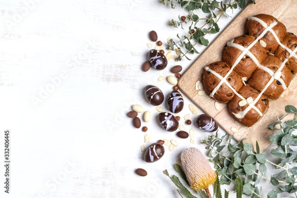 Fototapeta Chocolate Easter eggs and homemade hot cross buns on a wooden serve board surrounded by shaved almonds, hazelnuts plus Australian native Banksia and Eucalyptus leaves on a rustic white background.