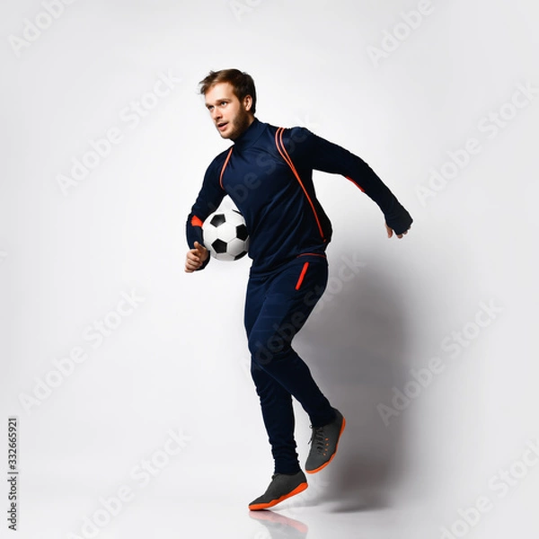 Fototapeta Soccer player in blue tracksuit, colored sneakers and backpack. He is running, holding ball, posing isolated on white Full-length