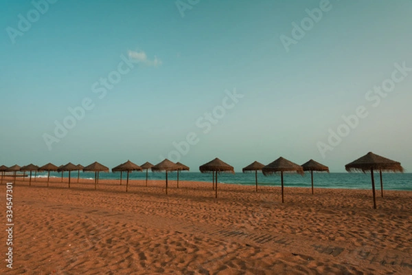 Fototapeta Empty sandy beach on Algarve coast with two rows of beach umbrellas on the background of blue sky. Tourism and travel concept.