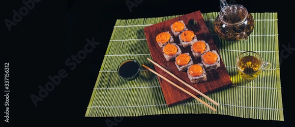 Fototapeta Delicious rolls on wooden board with chopsticks and green tea in teapot on decorative bamboo napkin. Close up of tasty sushi rolls on cutting board on black background.