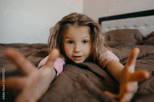 Fototapeta Child girl have fun  pajamas party during Covid-19 quarantine at home. Funny little girl  lying on the bed, shrug hands and surprise