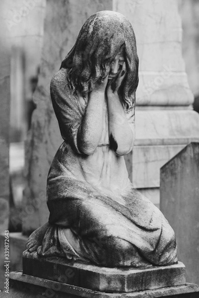 Fototapeta black and white sculpture of a angel girl in an old cemetery. Closeup of stoned angel with closed eyes and cross monument at cemetery. Graveyard old weathered stone sad baby statue on funeral.