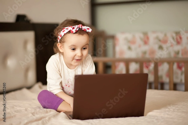 Fototapeta Little cute girl sitting on the bed and using a digital tablet laptop notebook. Online call friends or parents.