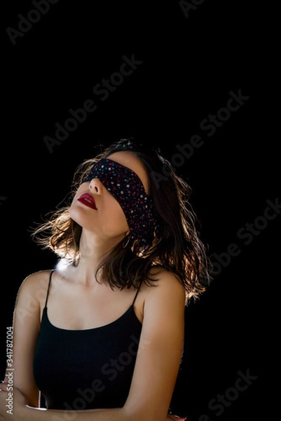 Fototapeta Portrait of beautiful young woman tying her eyes with black scarf on black background