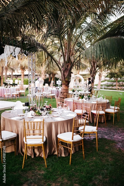 Fototapeta International Wedding outdoor celebration party under palm trees. Served tables on green area in hotel. Landyard. Beige and pink colors. Close-up and wide angle.