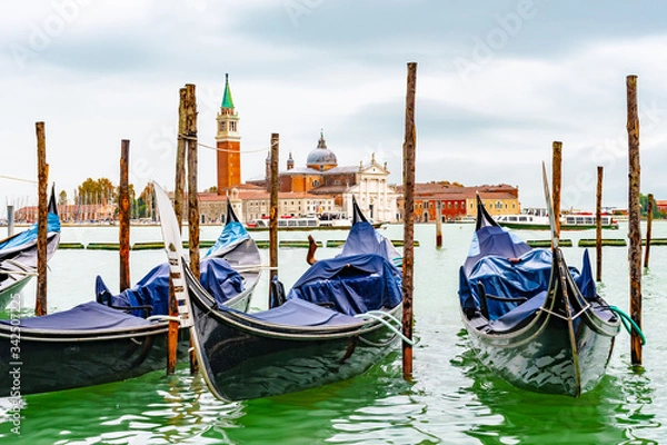 Fototapeta Venice, Italy. Empty Gondolas/ Gondole docked by lagoon mooring poles. Famous romantic city tour boat ride for tourists/ couples/ people. Bell Tower San Giorgio Maggiore Basilica Church in background