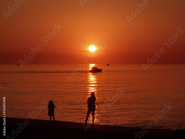 Fototapeta Mom and daughter watch beautiful sea sunset from the pier