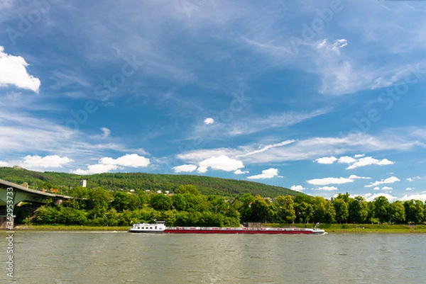 Fototapeta A barge flowing on the river Rhine in western Germany with a blue sky with clouds and a forest in the background.