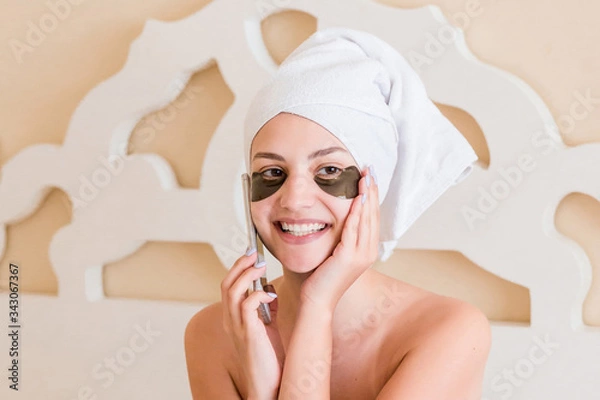 Fototapeta Smiling woman wrapped in towel after bath and with black eye patches is relaxing on the bed and talking on the phone. Beauty treatment concept